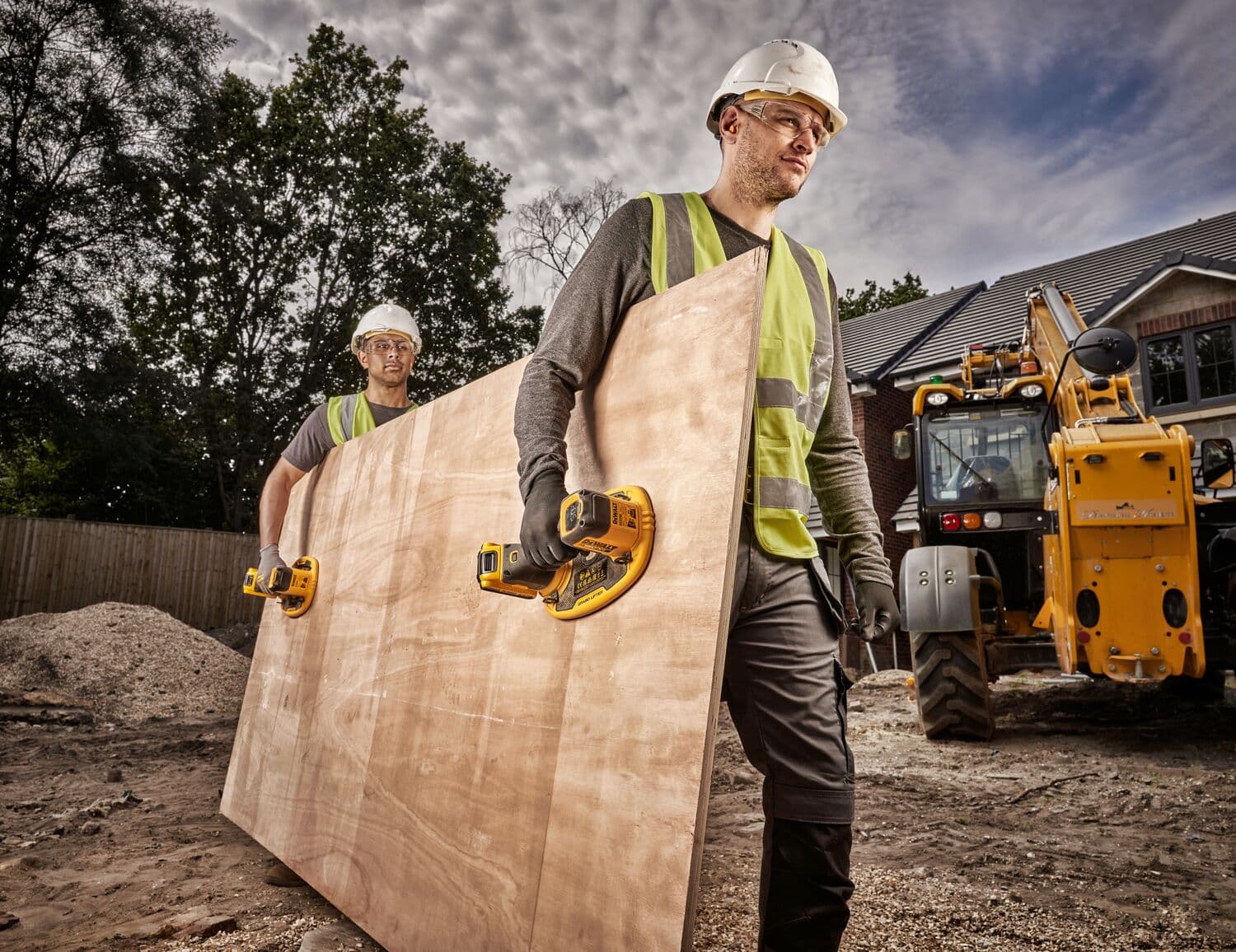 Two workers use DEWALT tools to carry a large wooden sheet at a construction site, with machinery in the background.
