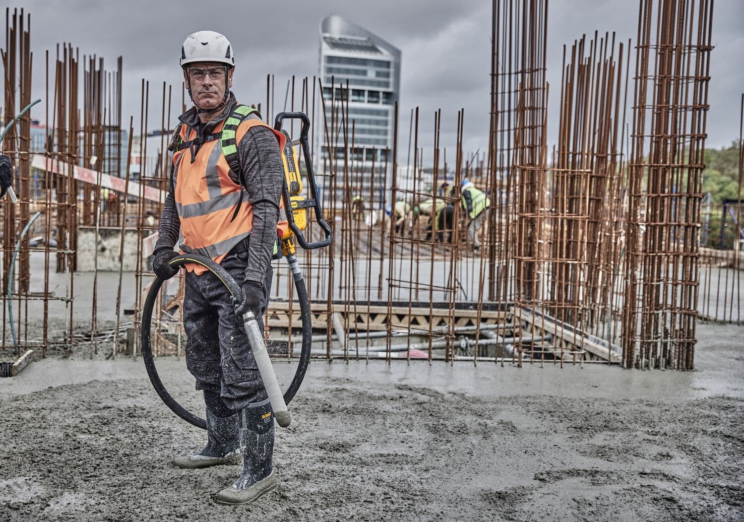 Person wearing a safety helmet and high-visibility vest, operating a DEWALT concrete vibrator tool (DCPS310N) at a construction site with rebar structures and wet concrete.