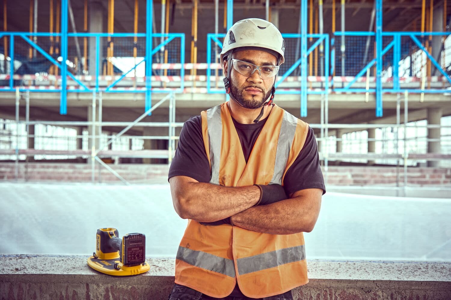 Worker in orange vest and helmet stands on site with DEWALT DCE590 tool on ledge in front.