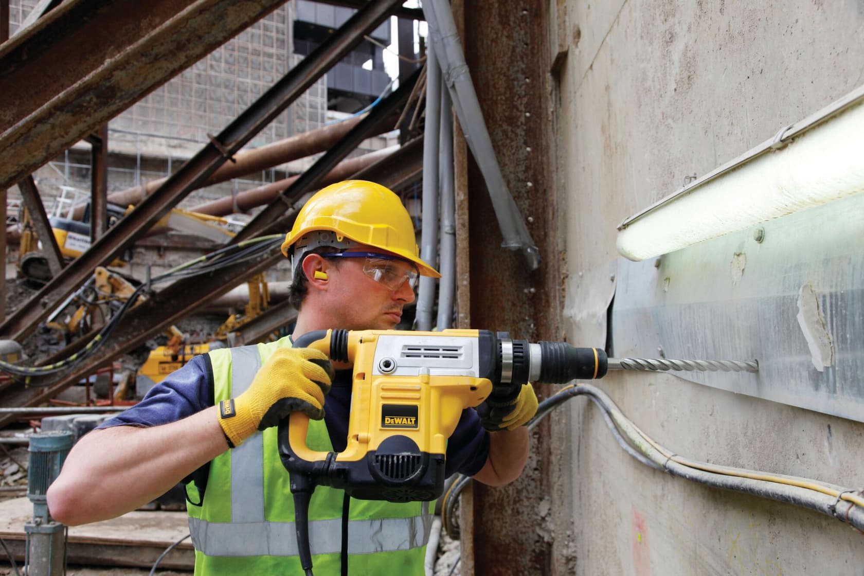 Person using a DEWALT rotary hammer drill on a construction site, wearing safety gear and drilling into a concrete wall.