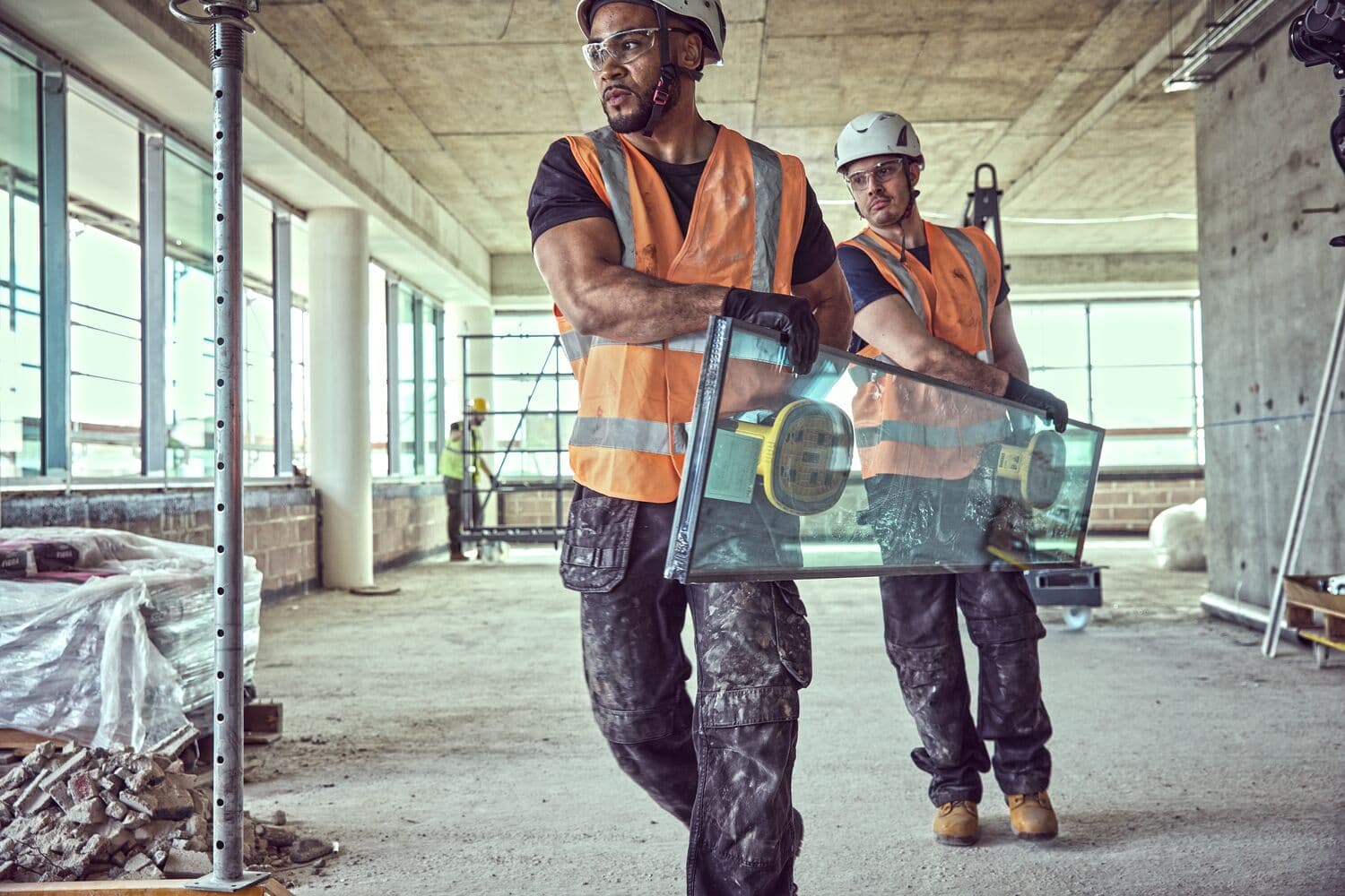 Two construction workers wearing orange safety vests and helmets are carrying a large glass panel with suction grips inside