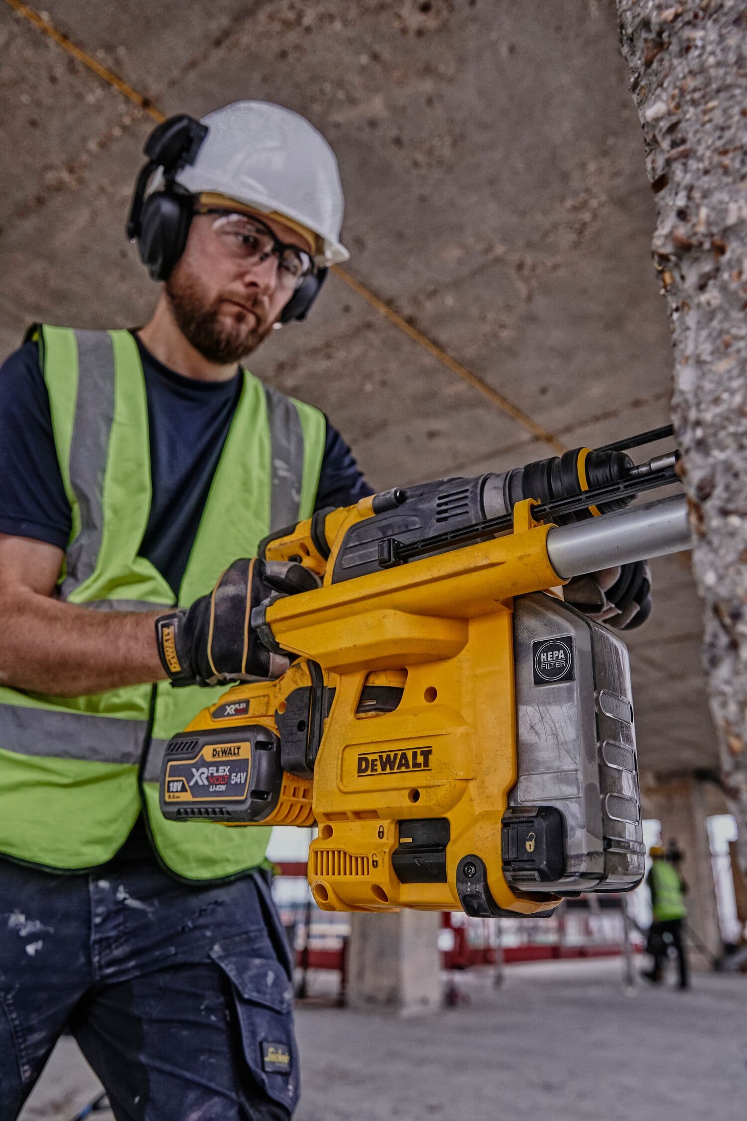 Worker holding a DEWALT DCH334X2 tool at a construction site, wearing safety gear and a green vest.