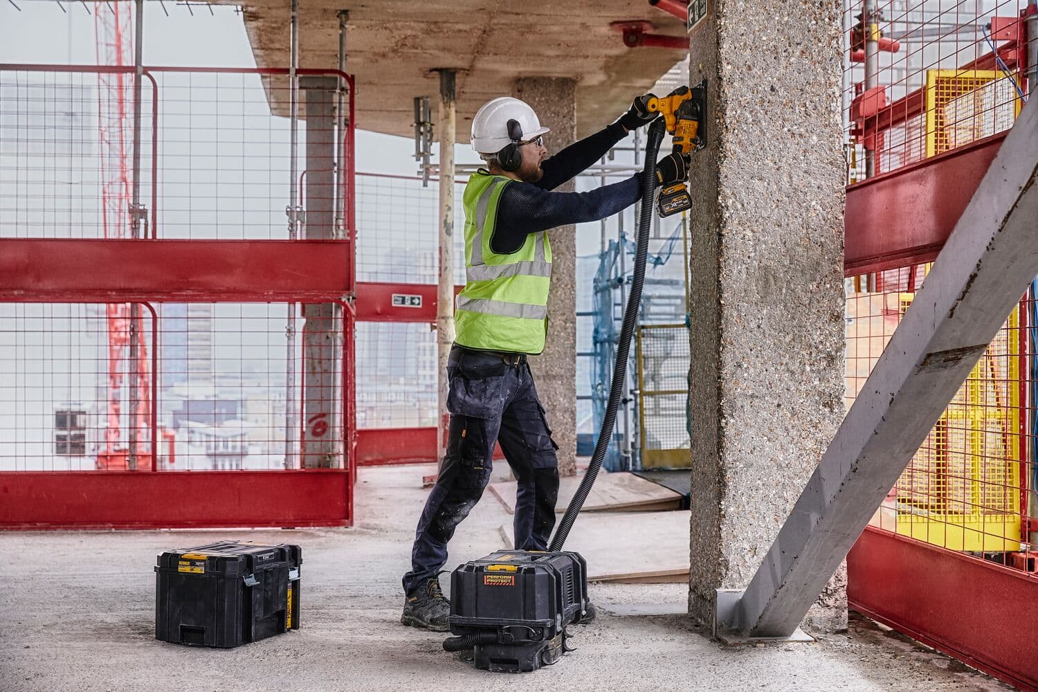 Worker uses DEWALT tool on concrete pillar at construction site, with DEWALT cases and safety gear visible.