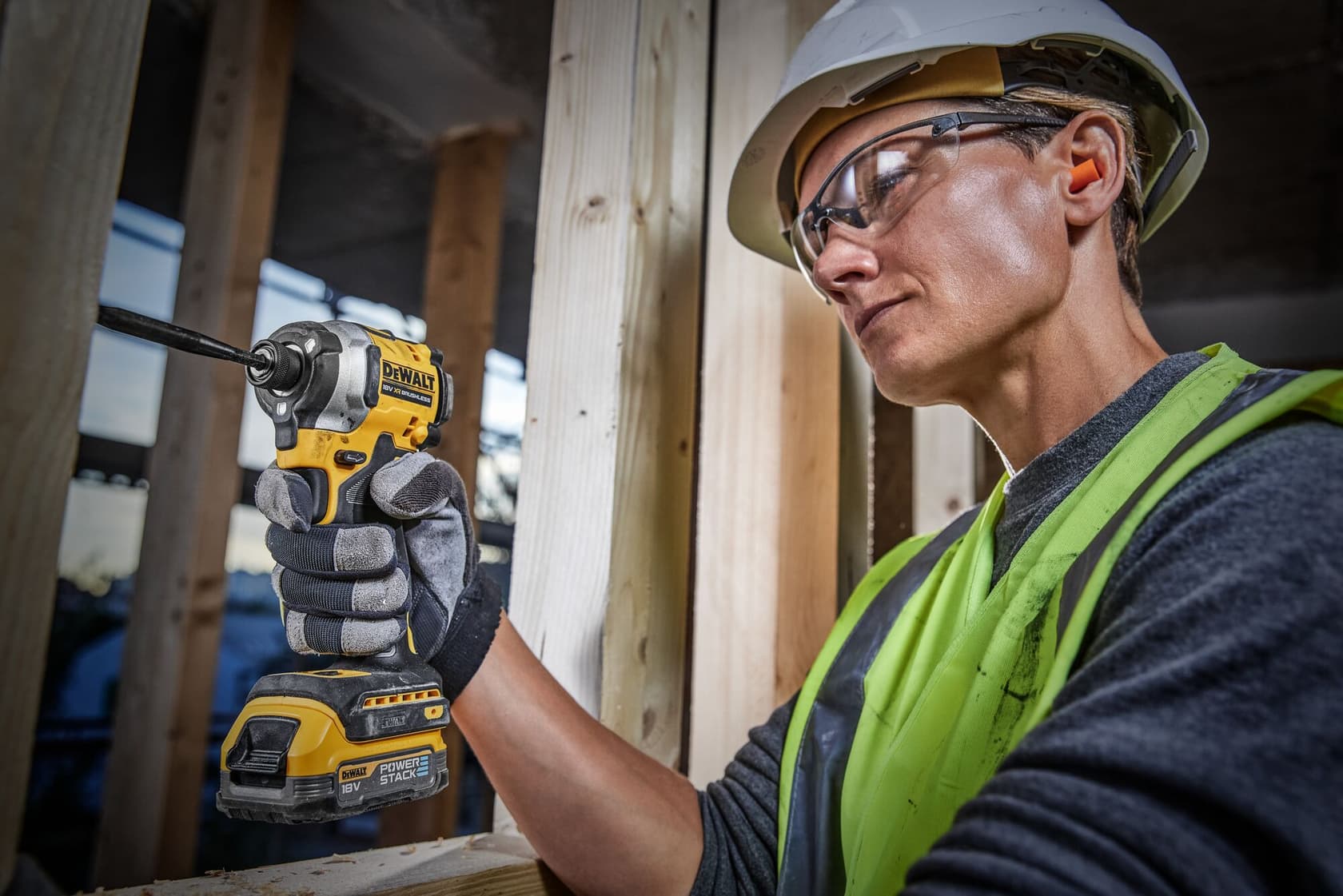 A person wearing a safety helmet, neon safety vest, and gloves uses a DEWALT DCF850E2T 18V cordless impact driver to work on