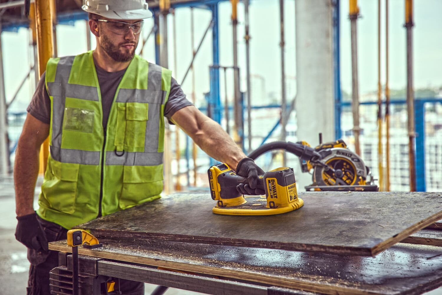 Worker uses a DEWALT DCE590 tool to sand a board at a construction site, wearing safety gear and gloves.