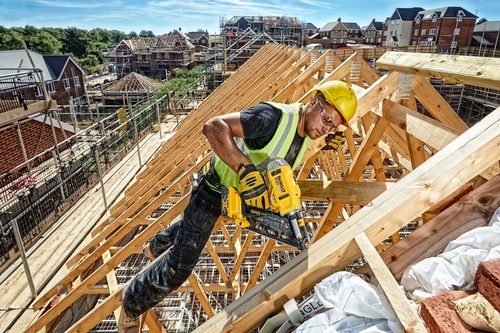 Worker uses a DEWALT nailer on wooden roof frame at a construction site with houses in the background.