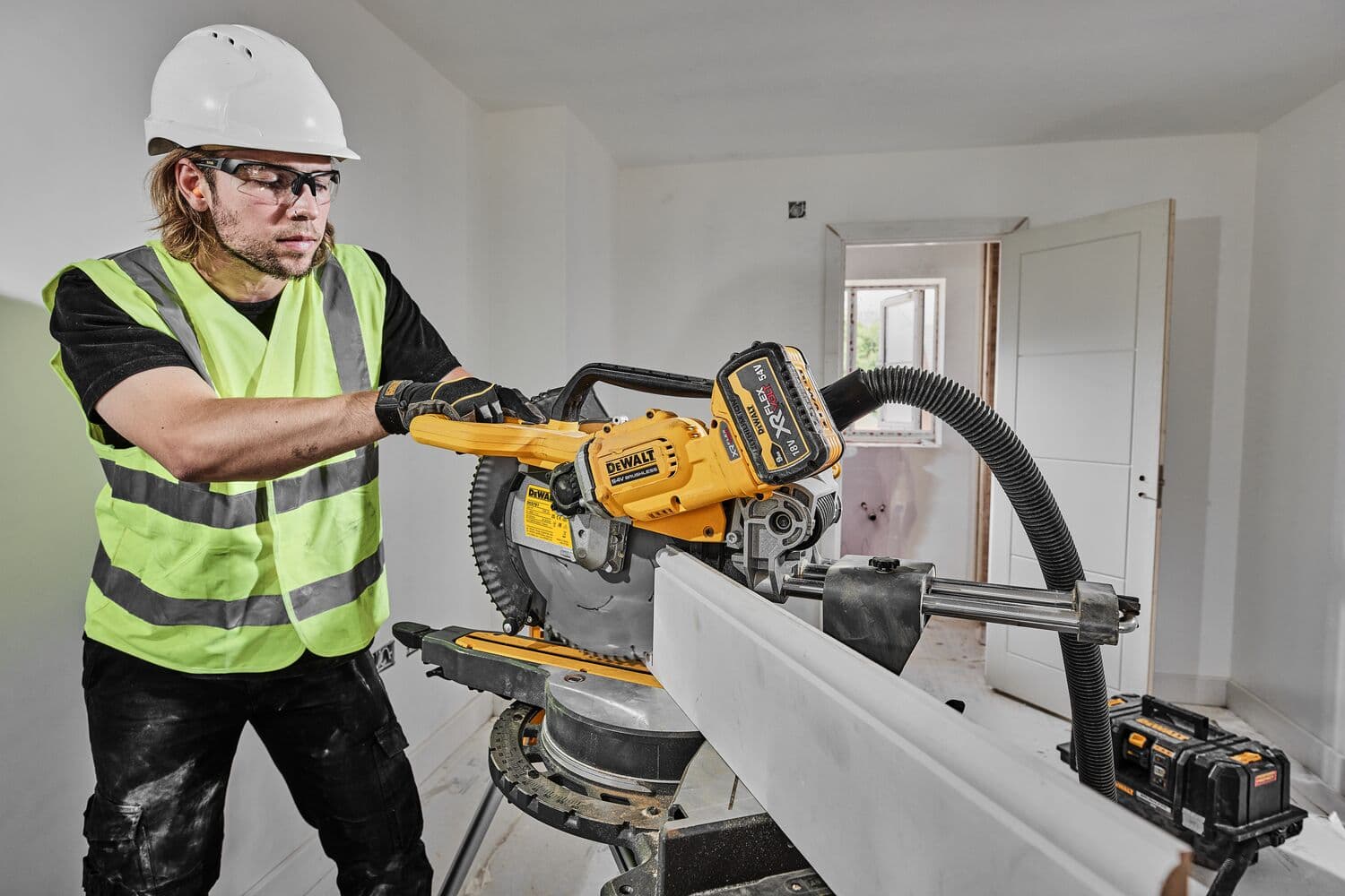 A construction worker wearing a safety helmet and high-visibility vest operates a DEWALT DCS781N cordless mitre saw to cut a