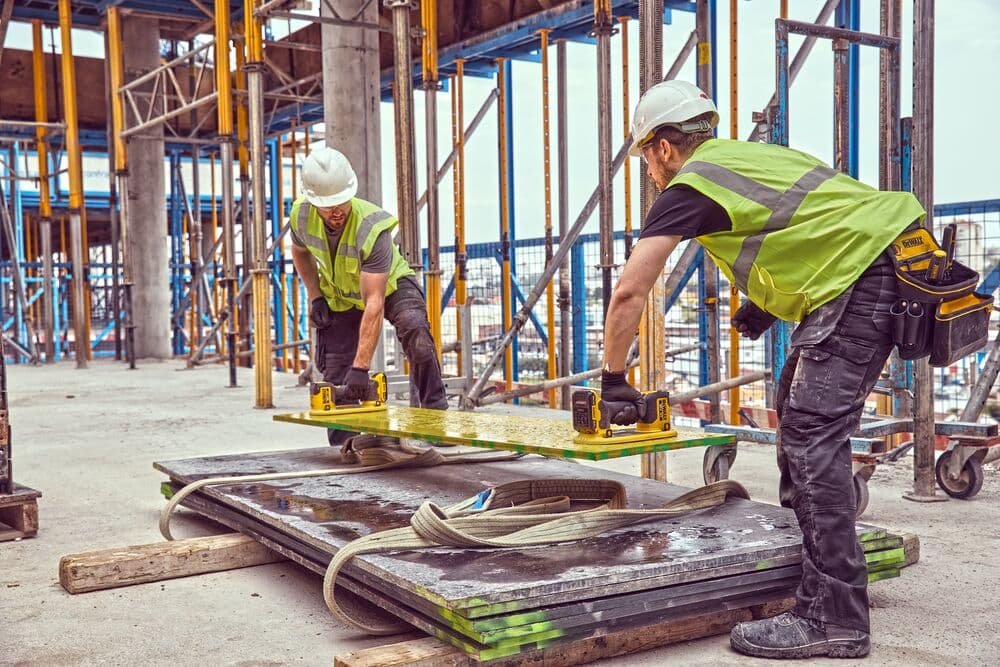 Two construction workers wearing high-visibility vests and hard hats are using DEWALT cordless tools to move a large metal