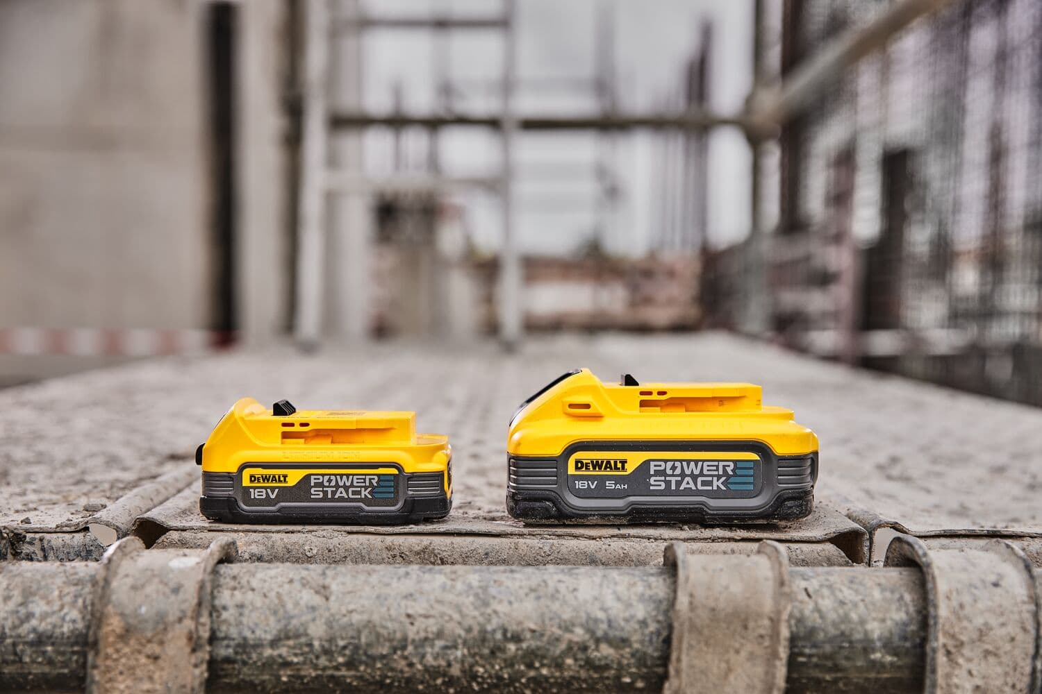 Two DEWALT POWERSTACK 18V batteries are shown on a construction site, placed side by side on a metal surface.