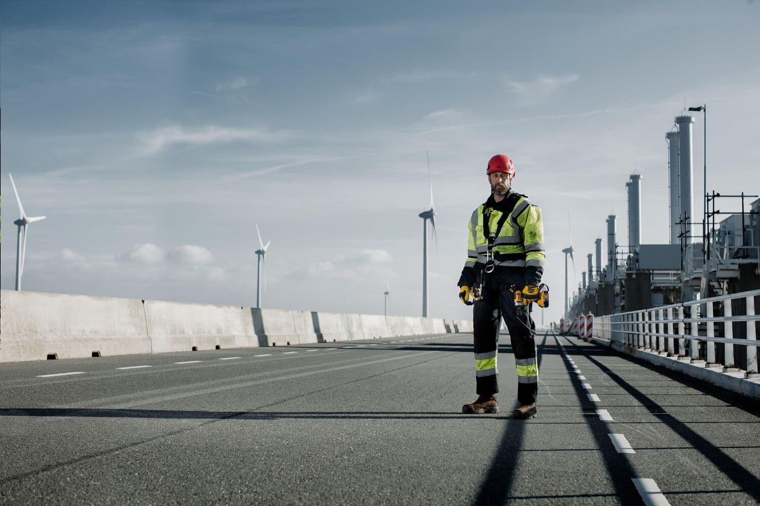 A construction worker wearing a high-visibility jacket and red helmet stands on an industrial road, holding DEWALT tools