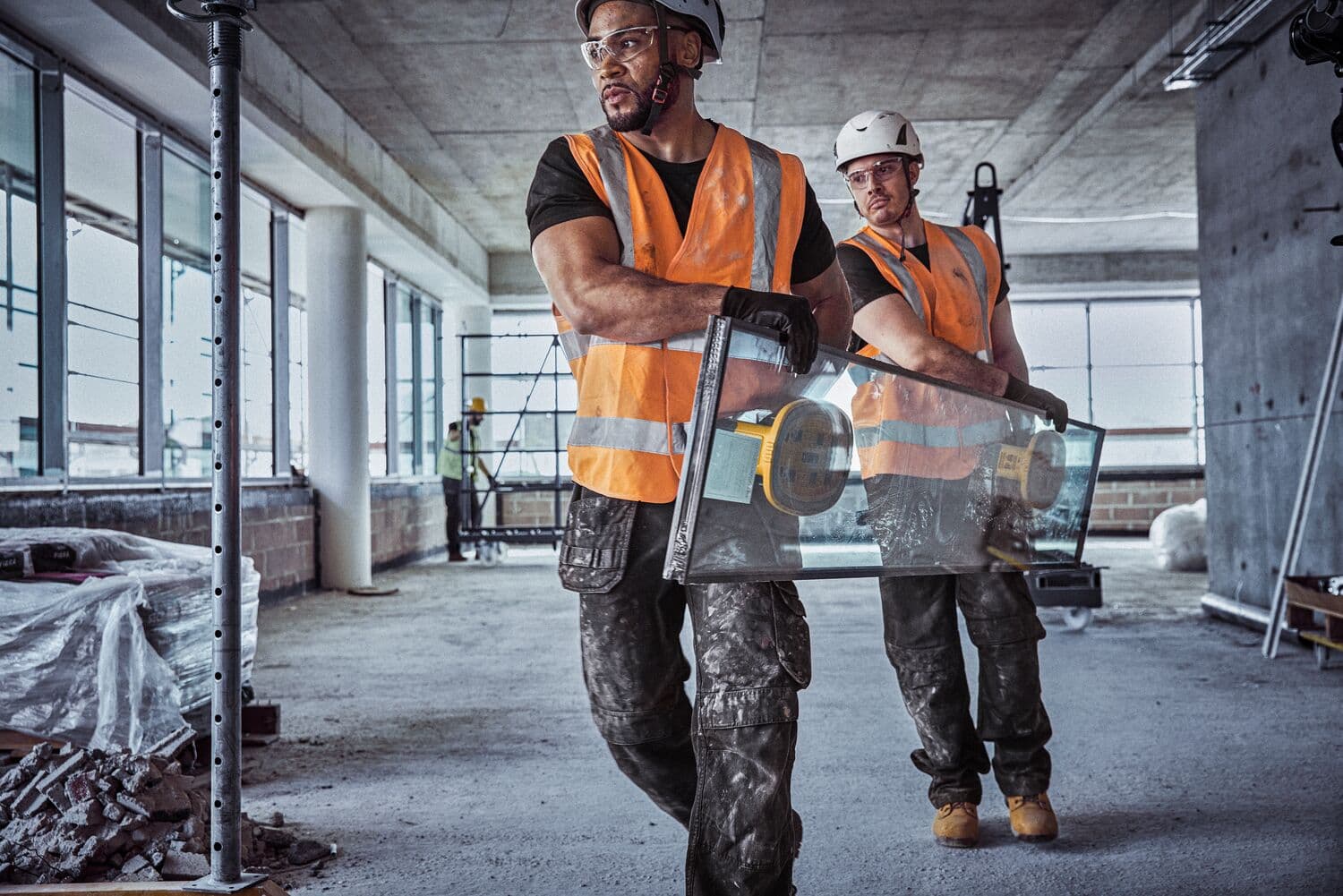 Two construction workers wearing orange safety vests and helmets are carrying a large glass sheet with a DEWALT suction cup tool inside a building under construction.
