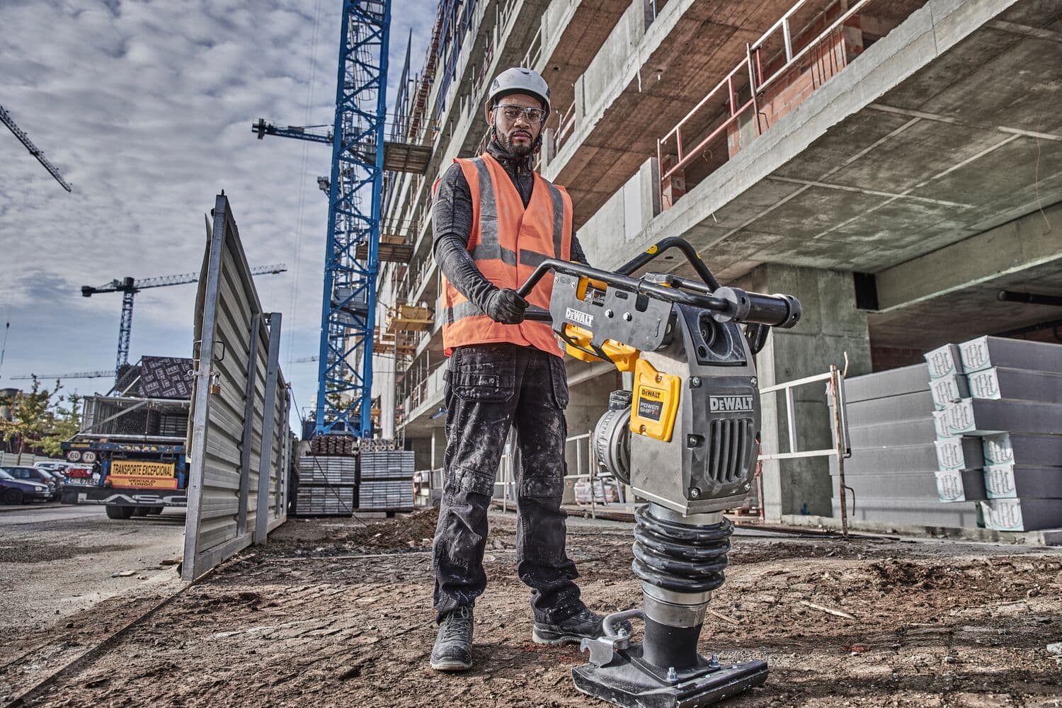 Person in safety gear using a DEWALT compactor at a construction site with cranes and a building in the background.