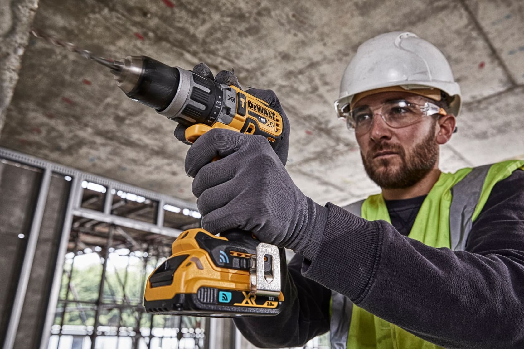 A person wearing safety gloves, a high-visibility vest, and a hard hat is using a DEWALT cordless drill at a construction site. The face is blurred for privacy.