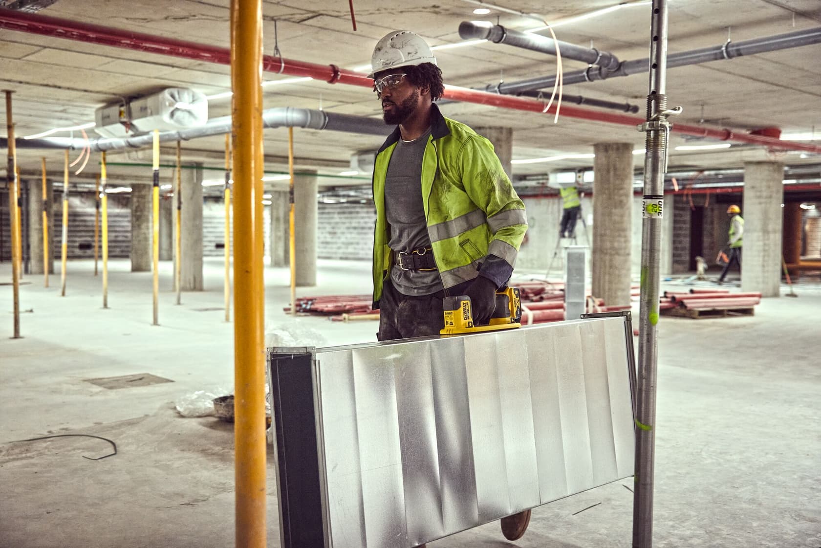 Worker wearing a hi-vis jacket and helmet carries metal sheet with a DEWALT tool in an unfinished construction site.