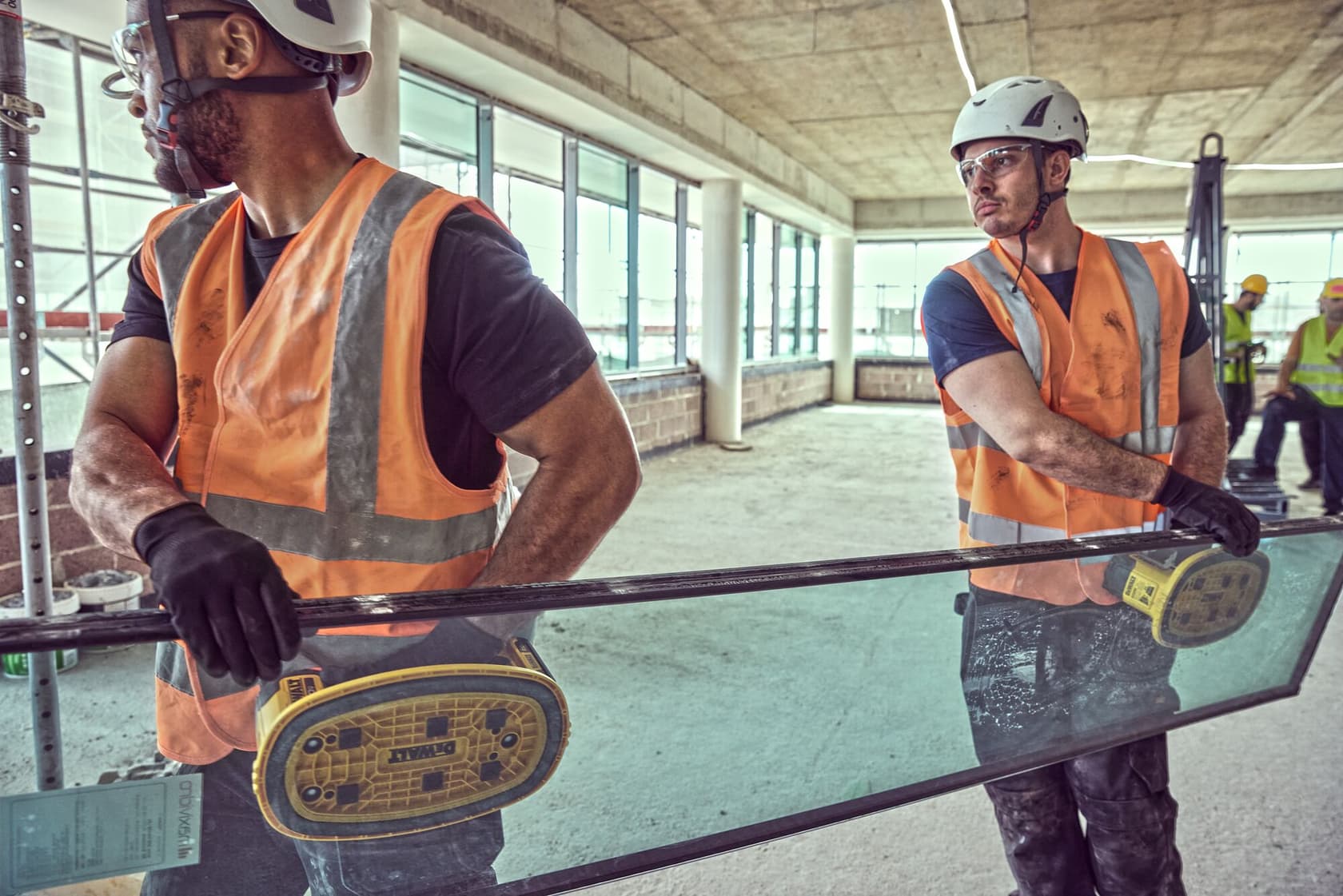 Two workers use DEWALT suction tools to carry glass indoors, both wearing safety gear and orange vests.