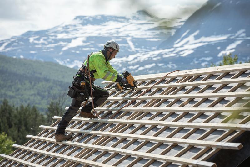 A worker wearing safety gear and a helmet uses a DEWALT nailer tool (DCN930N) on a wooden roof structure with mountain scenery in the background.