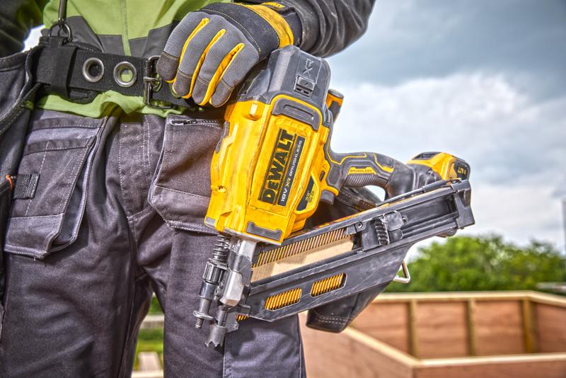 Close-up of a construction worker holding a DEWALT DCN930N cordless nailer. The worker is wearing protective gloves and a tool belt, with an outdoor construction site in the background.