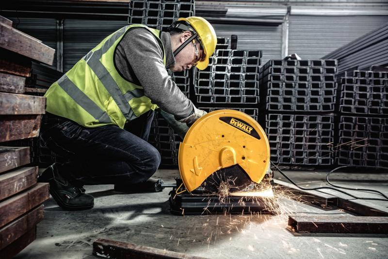 A worker wearing a high-visibility vest and helmet uses a DEWALT D28730 metal cutting chop saw to cut a metal bar, generating sparks in an industrial environment with stacks of metal beams.