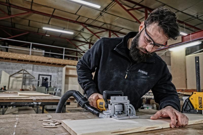 A person using a DEWALT DW682K woodworking tool on a piece of wood in a workshop. The person's face is blurred for privacy. The workspace features various woodworking equipment and wooden boards.