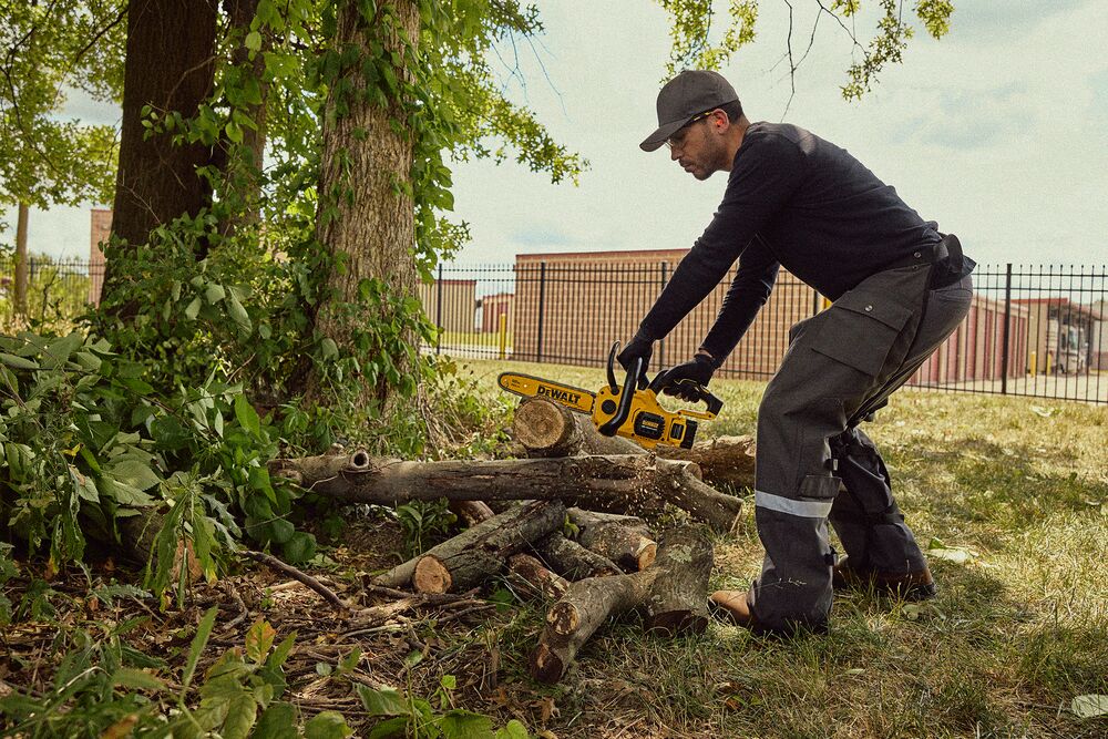 A person wearing protective clothing and gloves is using a yellow DEWALT cordless chainsaw to cut branches or logs outdoors near a tree.