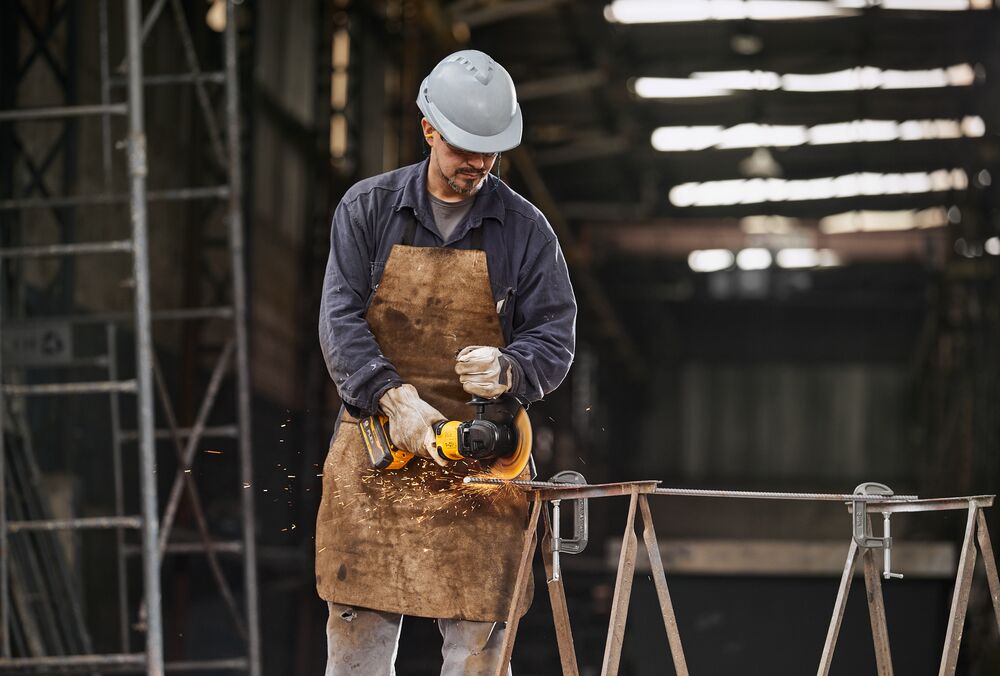A person wearing protective gear, including a helmet, gloves, and an apron, uses a DEWALT power tool to cut metal in an industrial workshop. Sparks are visible as the tool operates.
