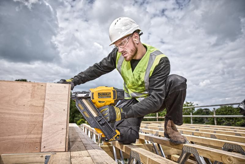 A construction worker wearing a safety helmet and high-visibility vest is using a yellow DEWALT DCN930N nailer to secure wooden boards at an outdoor building site. The worker's face is blurred for privacy.