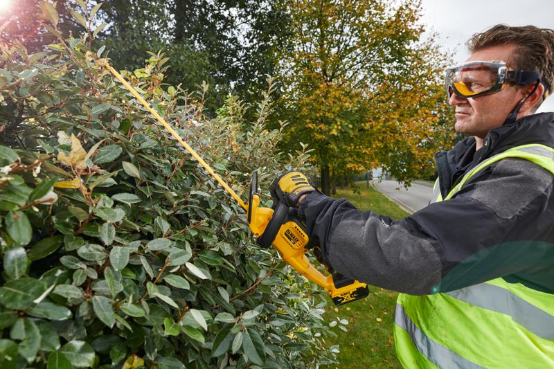 A person wearing safety gear and gloves is using a DEWALT DCM563PB cordless hedge trimmer to cut a dense bush outdoors. The person's face is blurred for privacy.
