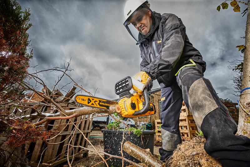 A person wearing safety gear uses a DEWALT DCM565N cordless brushless chainsaw to cut tree branches outdoors. The scene features cloudy skies, wooden pallets, and protective equipment.
