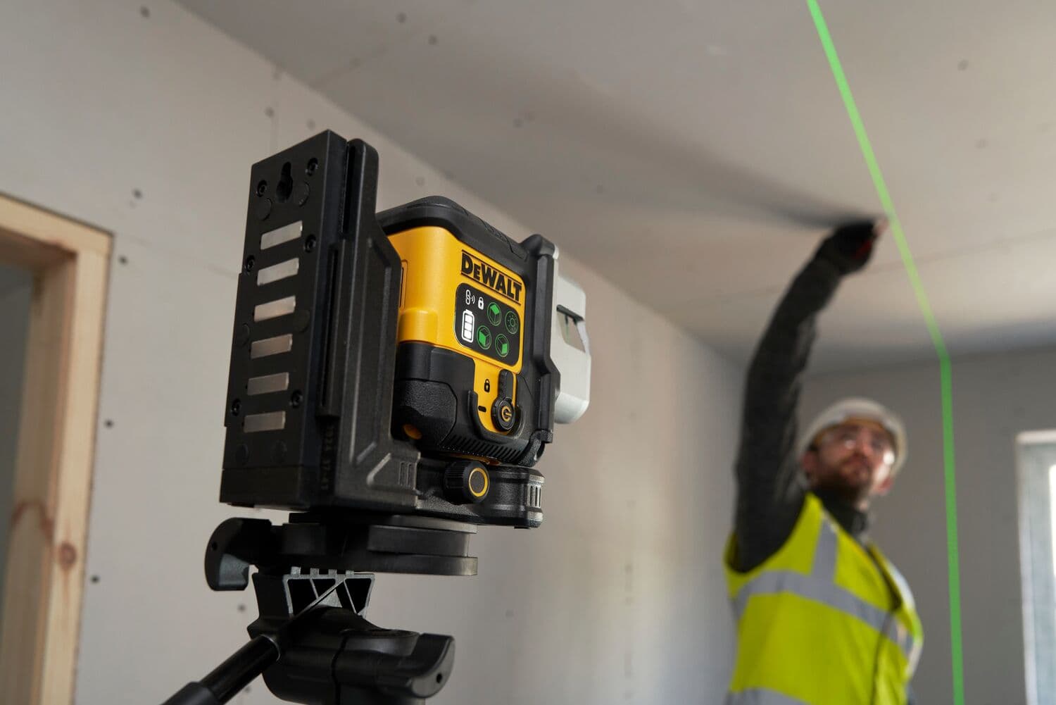 A DEWALT laser level device mounted on a tripod projects a bright green line onto a ceiling in a construction area. A person wearing a high-visibility vest is marking or measuring along the line.