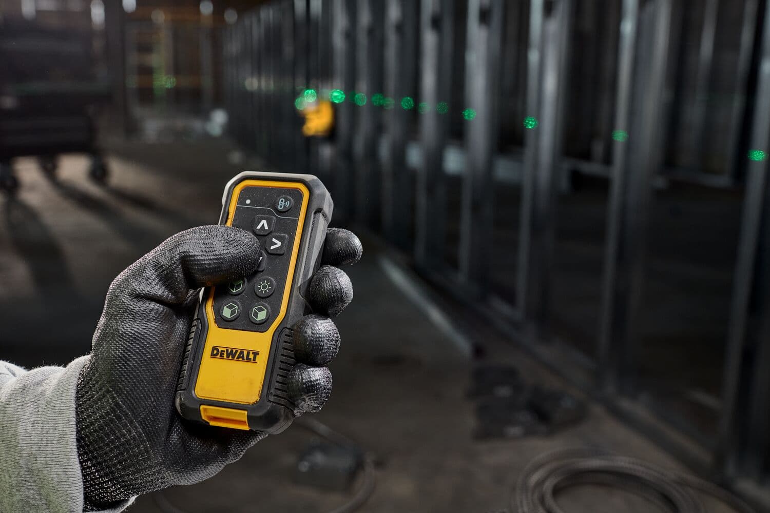 Gloved hand holding a DEWALT laser remote control, with green laser dots visible on a construction site.