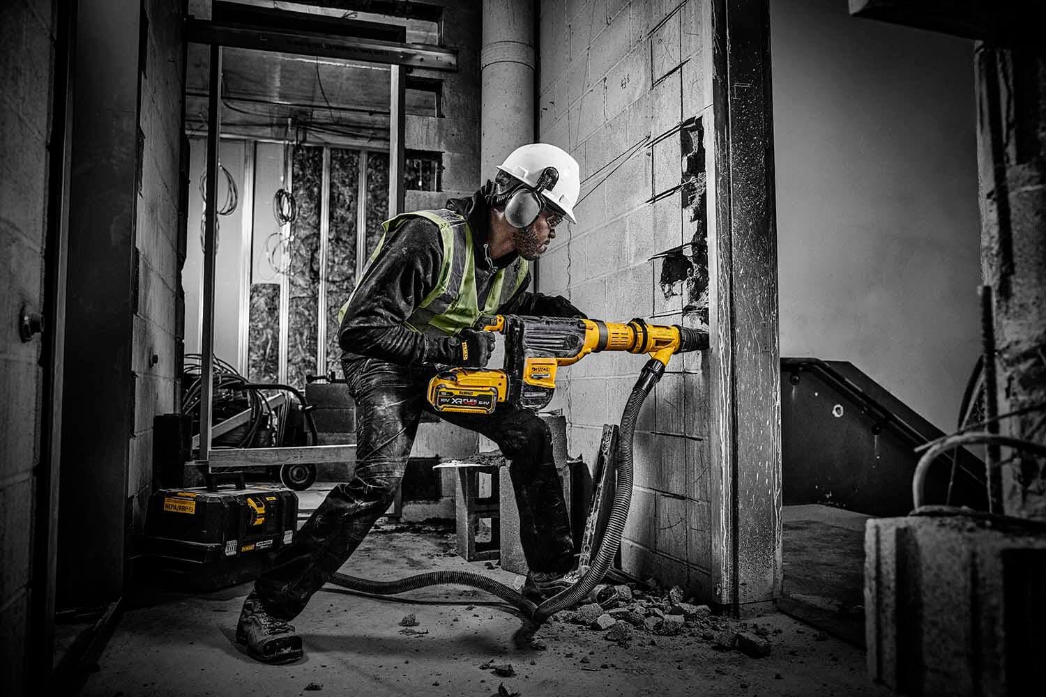 A worker wearing a safety helmet and vest operates a DEWALT DCH775 power tool to drill or demolish a concrete wall on a construction site. Debris is scattered on the floor, and the tool is connected to a hose.