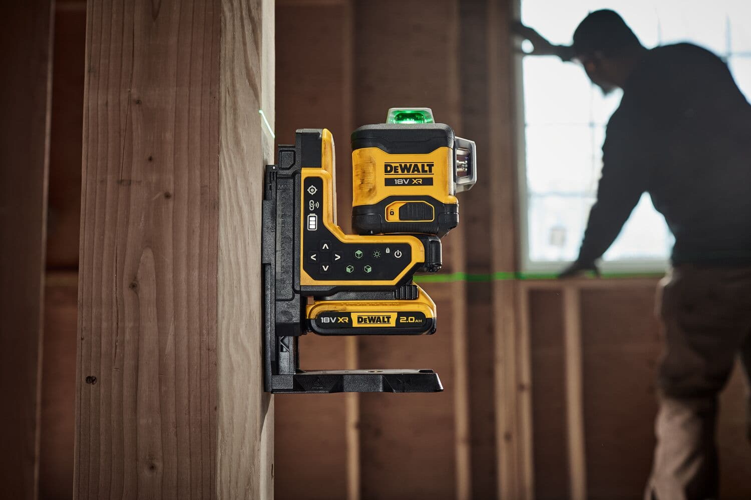 DEWALT laser level mounted on wood beam, projecting green lines, with person working in background.